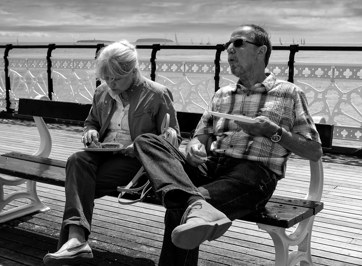 A man and a woman eating on a bench on a seaside pier