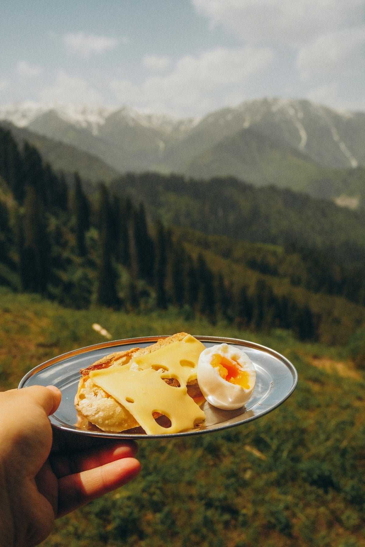 Hand holds breakfast plate with cheese and egg, mountains behind.
