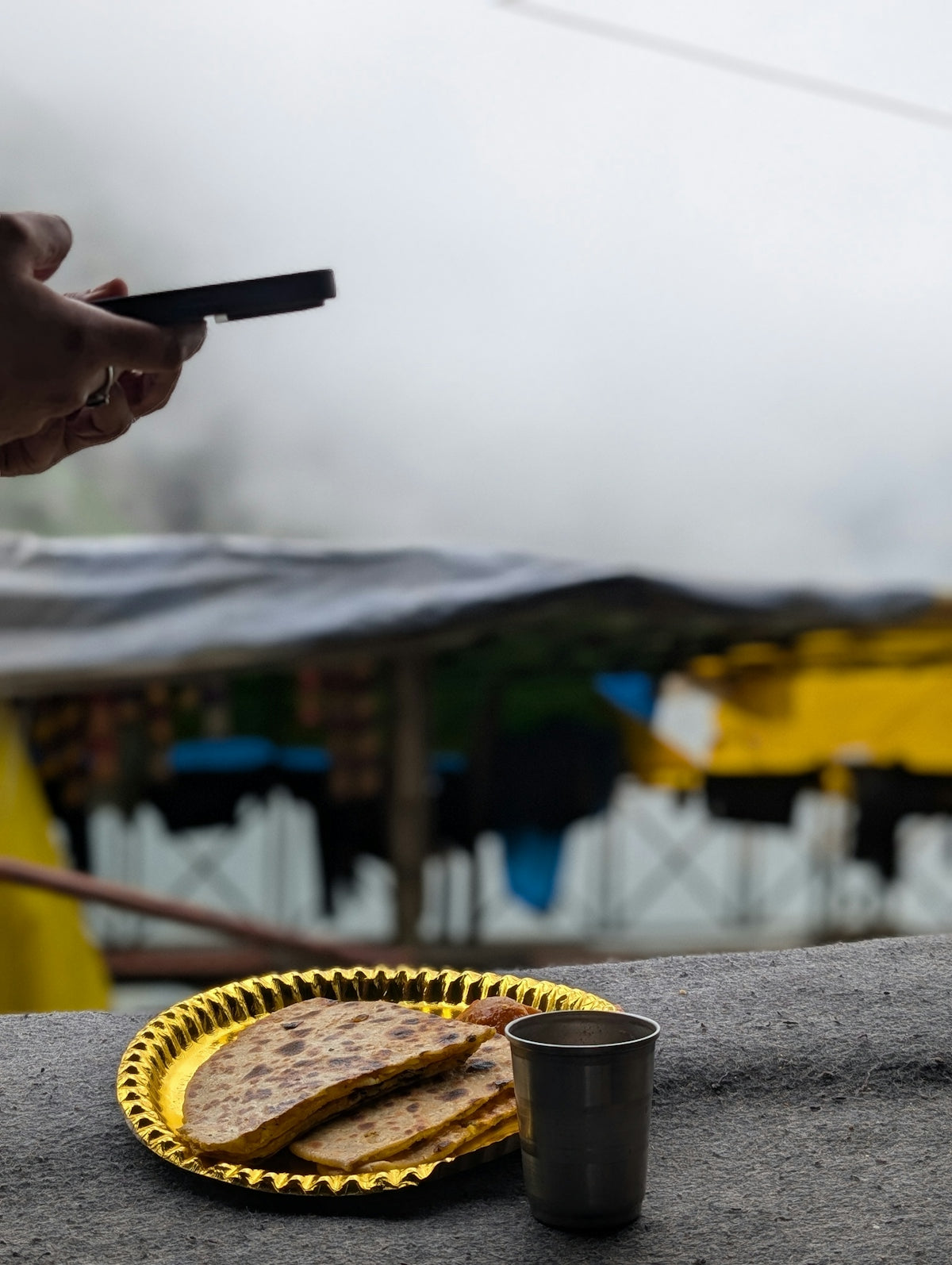 A person holding a phone over paratha and a metal cup on a ledge